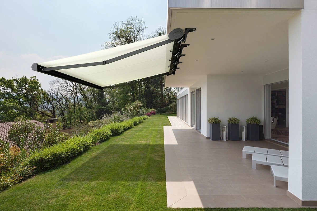 Duża biała markiza tarasowa zamontowana do elewacji nad przestronnym tarasem z płytkami i zielonym trawnikiem