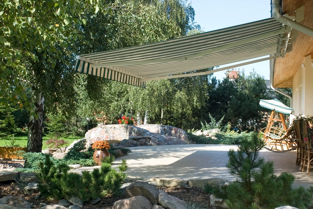 Taras z płytkami, ogrodowa aranżacja z kamiennym niskim murkiem i rozwiniętą markizą tarasową