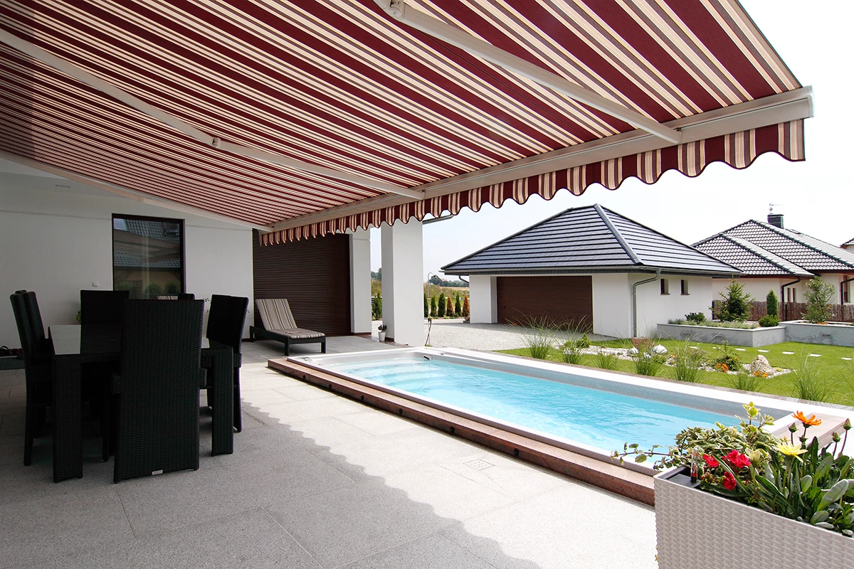 Taras z markizą tarasową w czerwono-białe pasy nad przestrzenią z meblami ogrodowymi i basenem przy domu