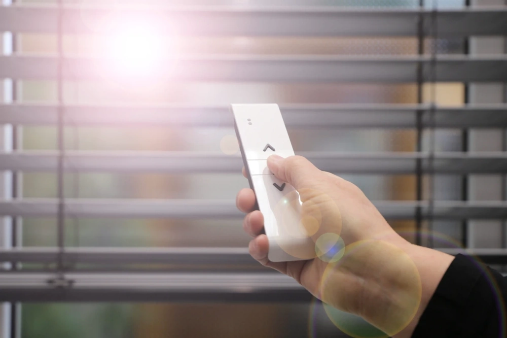 Ręka z pilotem steruje żaluzjami fasadowymi przed oknem